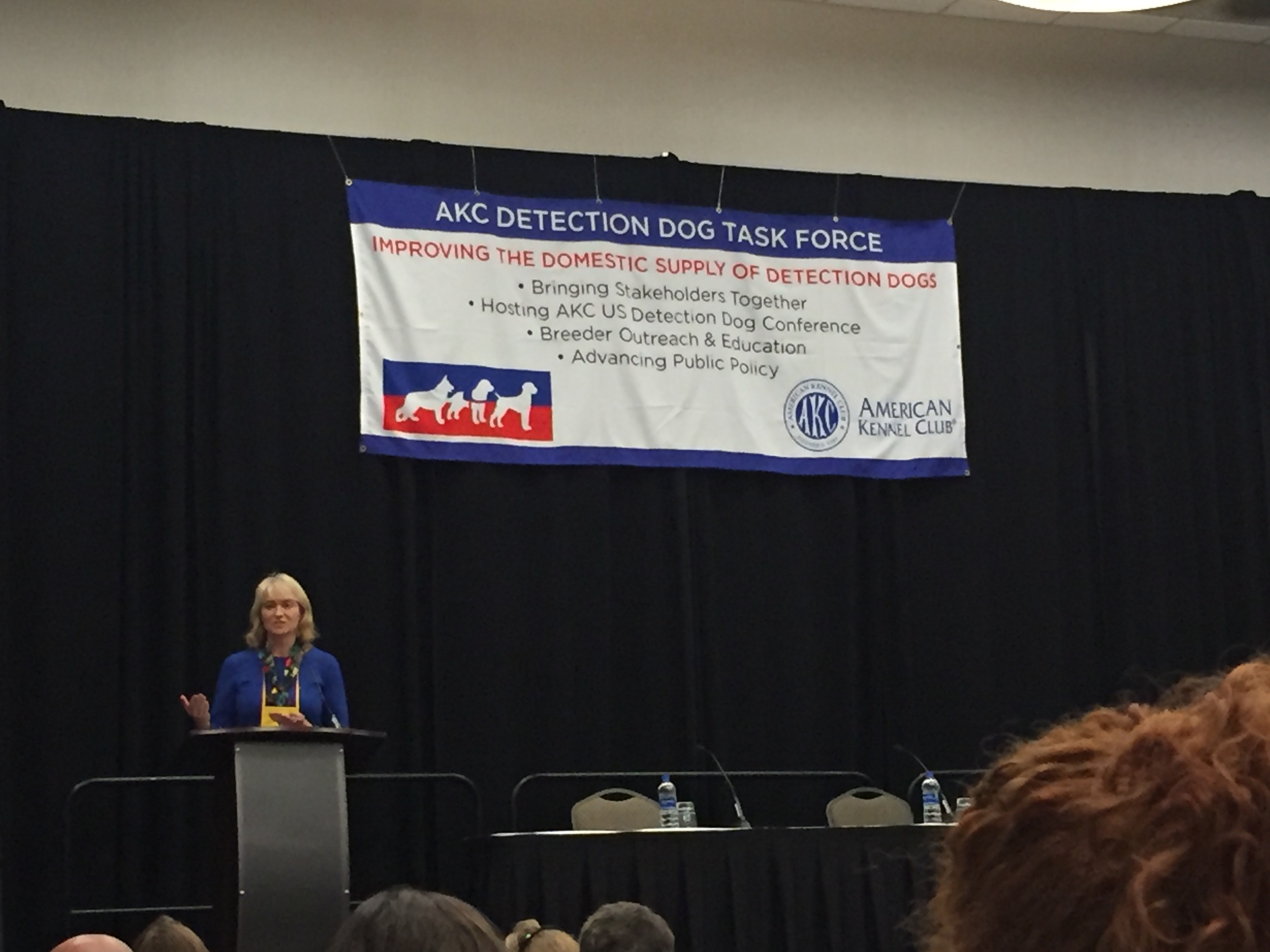 2019 AKC US Detection Dog Conference.  Sheila Goffe, Vice President, AKC Government Relations, speaking underneath a banner that explains the goals of the Conference.  Sheila is an insightful person who during her academic years earned a Fulbright Scholarship.
